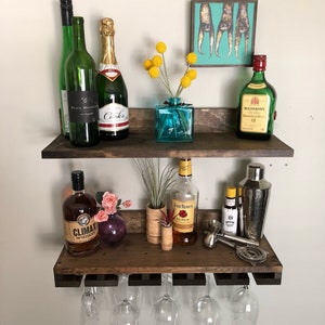 May include: A rustic, two-tiered wooden bar shelf with various bottles of liquor, champagne, and wine. The top shelf features a small turquoise vase with yellow flowers and a framed art piece. The bottom shelf holds glassware and bar tools.