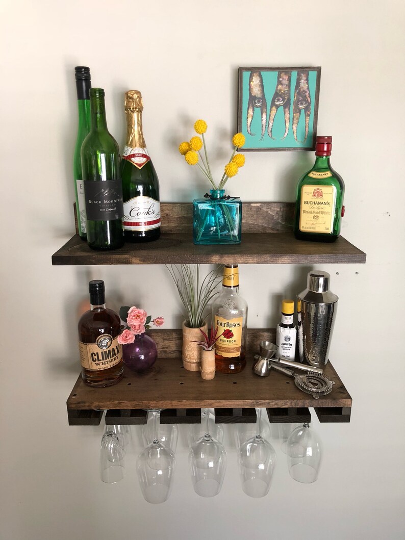 May include: A rustic, two-tiered wooden bar shelf with various bottles of liquor, champagne, and wine. The top shelf features a small turquoise vase with yellow flowers and a framed art piece. The bottom shelf holds glassware and bar tools.