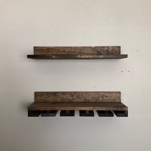 May include: Two-piece wooden wine rack set. The top shelf is a simple, rectangular design. The bottom shelf has slots for hanging wine glasses. Both shelves are stained in a dark brown color, with visible wood grain.