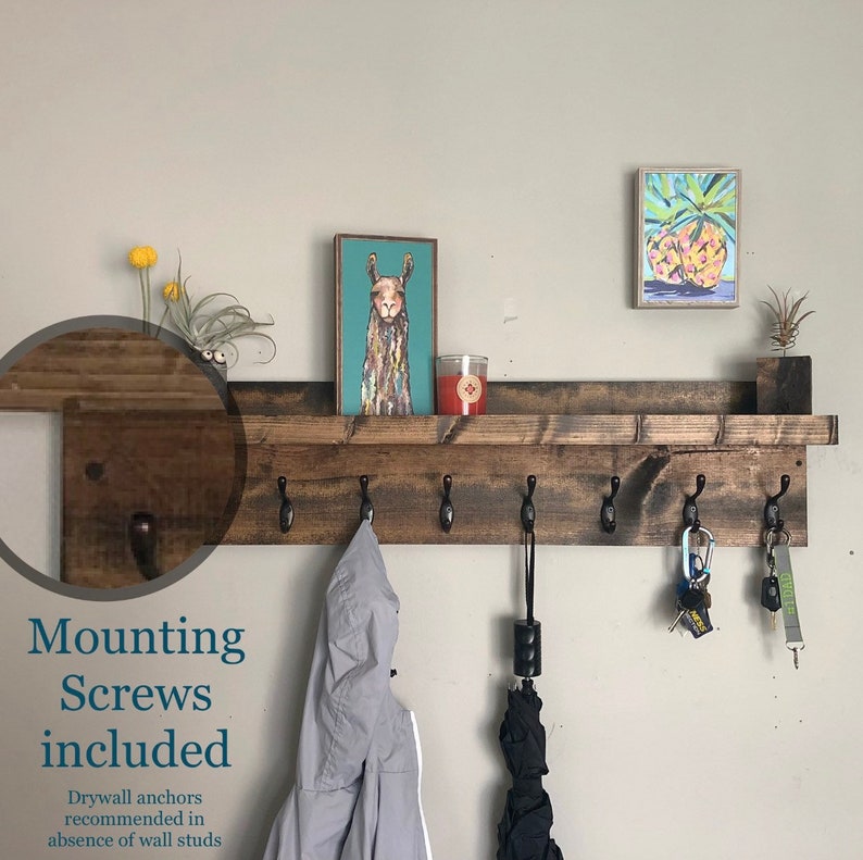 May include: A rustic wooden shelf with six hooks, a small shelf on top, and a dark brown stain. The shelf is mounted on a white wall and has a variety of items hanging from the hooks, including a jacket, an umbrella, and keys. The text "Mounting Screws included" is visible below the shelf.  Drywall anchors are recommended in the absence of wall studs.