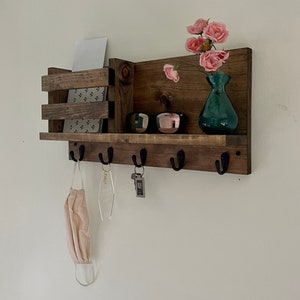 May include: A wall-mounted dark brown wooden entryway organizer featuring a horizontal shelf, a vertical mail slot on the left, and five black metal hooks along the bottom. The shelf holds a teal glass vase with pink flowers and sunglasses.