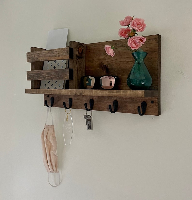 May include: A wall-mounted dark brown wooden entryway organizer featuring a horizontal shelf, a vertical mail slot on the left, and five black metal hooks along the bottom. The shelf holds a teal glass vase with pink flowers and sunglasses.