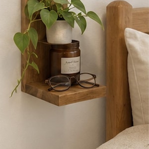 May include: A two-tiered wooden wall shelf holding a small potted green plant on the top level, and a brown glass jar with a white label and a pair of brown-rimmed eyeglasses on the bottom level, mounted on a plain wall next to a wooden bed frame.
