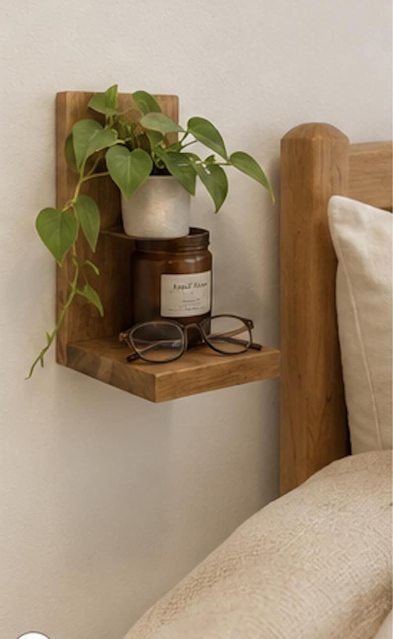 May include: A two-tiered wooden wall shelf holding a small potted green plant on the top level, and a brown glass jar with a white label and a pair of brown-rimmed eyeglasses on the bottom level, mounted on a plain wall next to a wooden bed frame.
