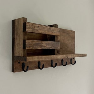 May include: A wall-mounted wooden organizer featuring a dark brown stained finish. The unit includes a rectangular shelf, a small storage compartment with two horizontal slats, and five black metal hooks attached to the bottom front panel.