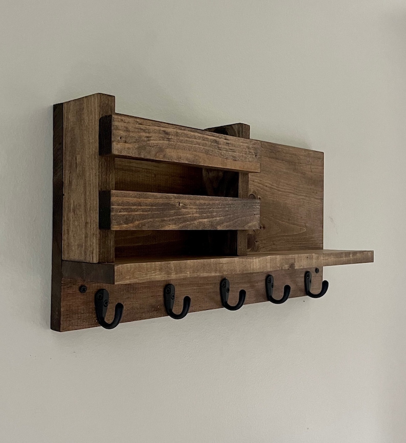 May include: A wall-mounted wooden organizer featuring a dark brown stained finish. The unit includes a rectangular shelf, a small storage compartment with two horizontal slats, and five black metal hooks attached to the bottom front panel.
