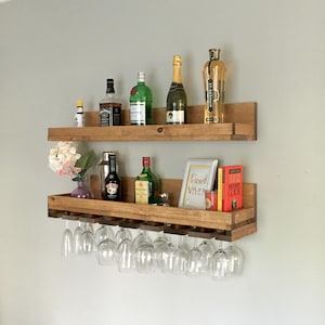 May include: Two wooden shelves with a wine glass rack underneath. The shelves are mounted on a wall and hold various bottles of liquor, a shaker, and a few decorative items. The top shelf has a bottle of St. Germain liqueur, a bottle of champagne, and a bottle of green liquor. The bottom shelf has a bottle of whiskey, a bottle of green liquor, a shaker, and a small decorative item. The wine glass rack holds 10 wine glasses.