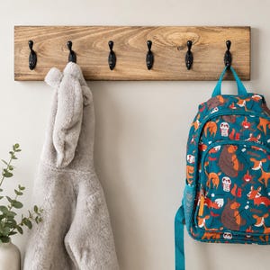May include: A rectangular wooden wall rack with six black metal hooks. A gray faux fur hooded jacket hangs on the left, and a teal backpack with a repeating pattern of bears, foxes, and campfires hangs on the right side of the wooden rack.
