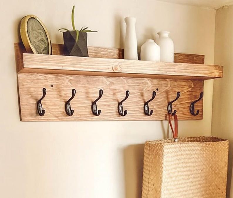 Puede incluir: Estante de pared de madera con seis ganchos de metal negro. El estante tiene un acabado de madera natural y un peque&ntilde;o estante superior que contiene objetos decorativos, incluyendo un marco de fotos, una planta y jarrones blancos. Una cesta tejida cuelga debajo.