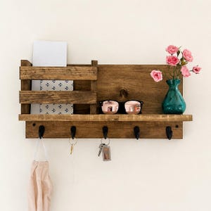 May include: A wall-mounted wooden organizer featuring a slatted mail slot, a flat shelf, and five black metal hooks. The shelf holds a teal glass vase with pink flowers and black sunglasses. A beige fabric mask hangs from one of the hooks.