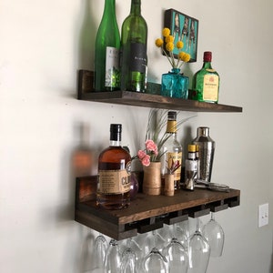 May include: A dark stained wooden wall-mounted bar shelf with two levels. The top shelf holds green wine bottles, a small blue vase with yellow flowers, and a liquor bottle. The bottom shelf has liquor bottles, a cocktail shaker, and wine glasses.