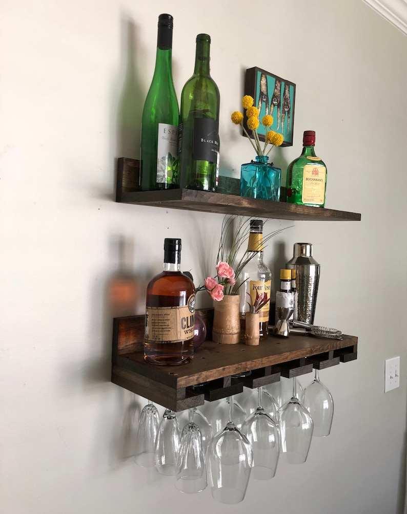 May include: A dark stained wooden wall-mounted bar shelf with two levels. The top shelf holds green wine bottles, a small blue vase with yellow flowers, and a liquor bottle. The bottom shelf has liquor bottles, a cocktail shaker, and wine glasses.
