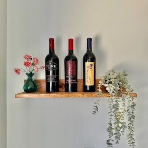 May include: Three bottles of red wine on a wooden shelf. The bottles are labeled "Cline", "Apothic Red", and "Blackstone". A small vase with pink flowers and a potted plant are also on the shelf.
