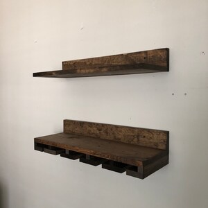 May include: Two rustic, dark-stained wooden shelves mounted on a white wall. The top shelf is a simple, rectangular design. The bottom shelf features cutouts for holding wine glasses, creating a functional and decorative storage solution.