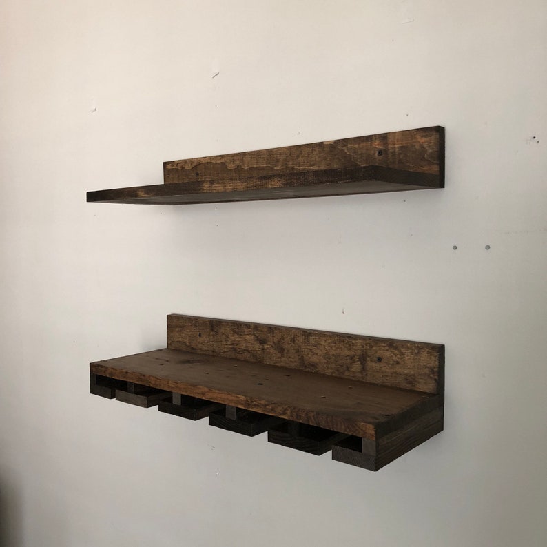 May include: Two rustic, dark-stained wooden shelves mounted on a white wall. The top shelf is a simple, rectangular design. The bottom shelf features cutouts for holding wine glasses, creating a functional and decorative storage solution.