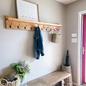 Puede incluir: Una estanter&iacute;a de pared de madera con ganchos negros, que sostiene una chaqueta vaquera y una cesta de flores tejida. Un letrero enmarcado con texto cuelga encima de la estanter&iacute;a. Un banco y cestas adicionales est&aacute;n debajo.