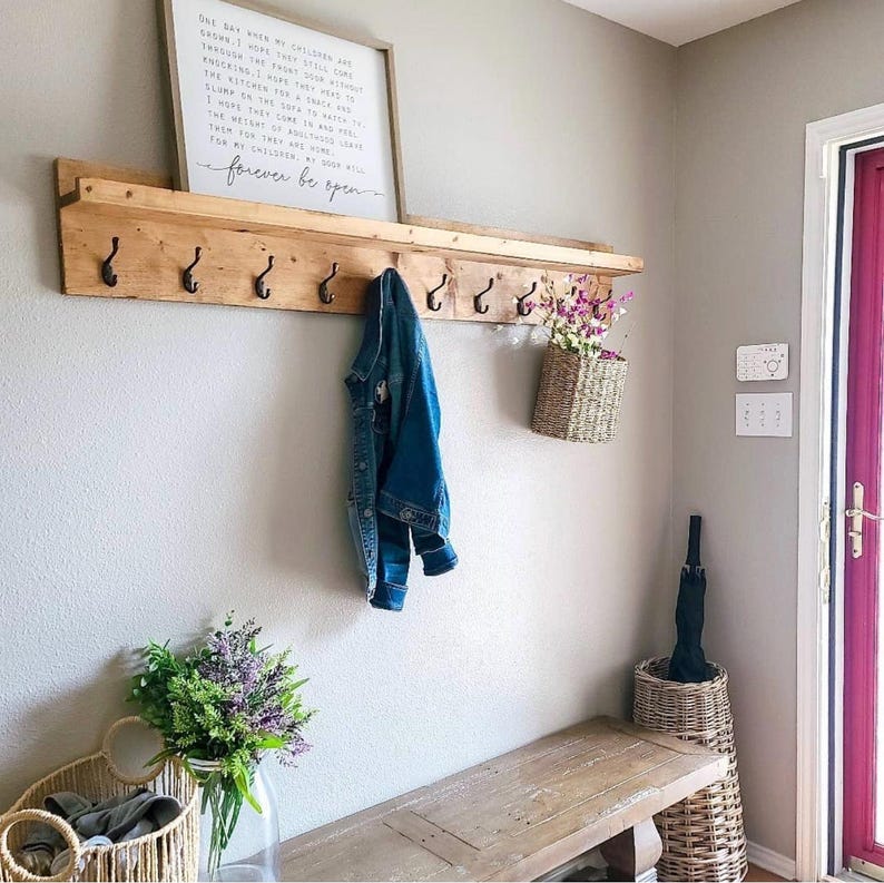 Puede incluir: Una estanter&iacute;a de pared de madera con ganchos negros, que sostiene una chaqueta vaquera y una cesta de flores tejida. Un letrero enmarcado con texto cuelga encima de la estanter&iacute;a. Un banco y cestas adicionales est&aacute;n debajo.