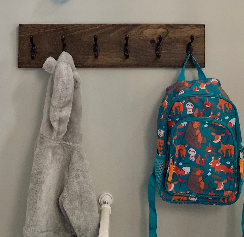 May include: A wooden coat rack with five hooks, a gray hooded sweatshirt hanging on one hook, and a teal blue backpack with a colorful animal print hanging on another hook.