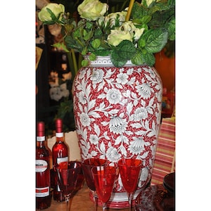 Könnte beinhalten: Eine rote und weiße Keramikvase mit einem Blumenmuster. Die Vase ist mit weißen Blumen und Blättern auf rotem Hintergrund verziert. Die Vase steht auf einem Tisch mit zwei Weinflaschen und vier Weingläsern.