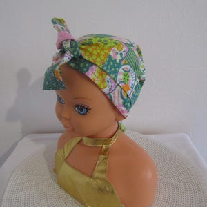 Peut inclure: Un foulard à motifs colorés dans les tons de vert, rose et jaune, noué sur le dessus. Le foulard est présenté sur une tête de mannequin aux yeux bleus et un haut doré. Le fond est un mur blanc uni.