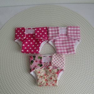 Peut inclure: Trois couches pour bébé en tissu rose et blanc avec différents motifs. Les couches sont en coton et ont une ceinture élastique blanche. Les motifs comprennent des cœurs roses, du vichy rose et blanc et un imprimé floral.