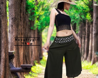 May include: A woman wearing a black crop top with lace detailing and a wide-leg, dark green, flowy pant with a white and green patterned belt. The woman is wearing a straw hat and is standing in a wooded area.