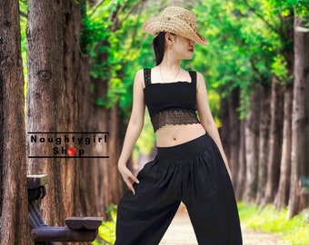 May include: A woman wearing a black crop top with lace trim and black harem pants. She is wearing a straw cowboy hat and is standing in a wooded area.