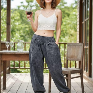 Könnte beinhalten: Eine Frau trägt ein weißes Spitzen-Crop-Top, eine dunkelgraue Haremshose und braune Sandalen. Sie trägt einen Strohhut und eine Sonnenbrille und hält ein Glas Rotwein. Die Szene spielt auf einer Außenterrasse mit Holzmöbeln.
