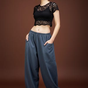 May include: A black lace crop top paired with loose-fitting, slate blue pants. The pants have large pockets and a relaxed silhouette. The model is standing in front of a brown background.