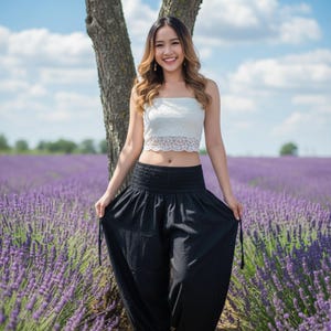 May include: A woman wearing a white lace crop top and black harem pants stands in a lavender field. The pants have a gathered waistband and drawstring ties. The sky is blue with fluffy white clouds.
