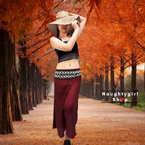 May include: A woman wearing a black lace crop top, a burgundy maxi skirt with a white and black patterned waistband, and a straw sun hat. The background features a path lined with trees with orange leaves.