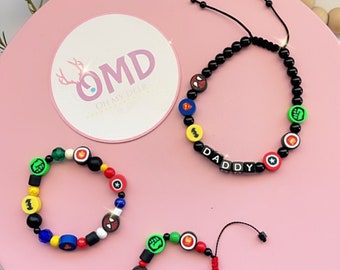 May include: Three colorful beaded bracelets with superhero logos and the word "DADDY" spelled out on one bracelet. The bracelets are on a pink surface with a white circle with the text "OMD OH MY DEER" in the center.
