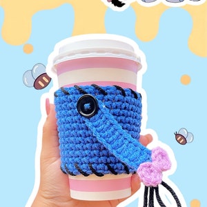 May include: A blue crocheted coffee cup sleeve with a black button and a pink tassel, held in a hand. The sleeve is wrapped around a pink and white striped cup. The image includes Winnie the Pooh characters and text.