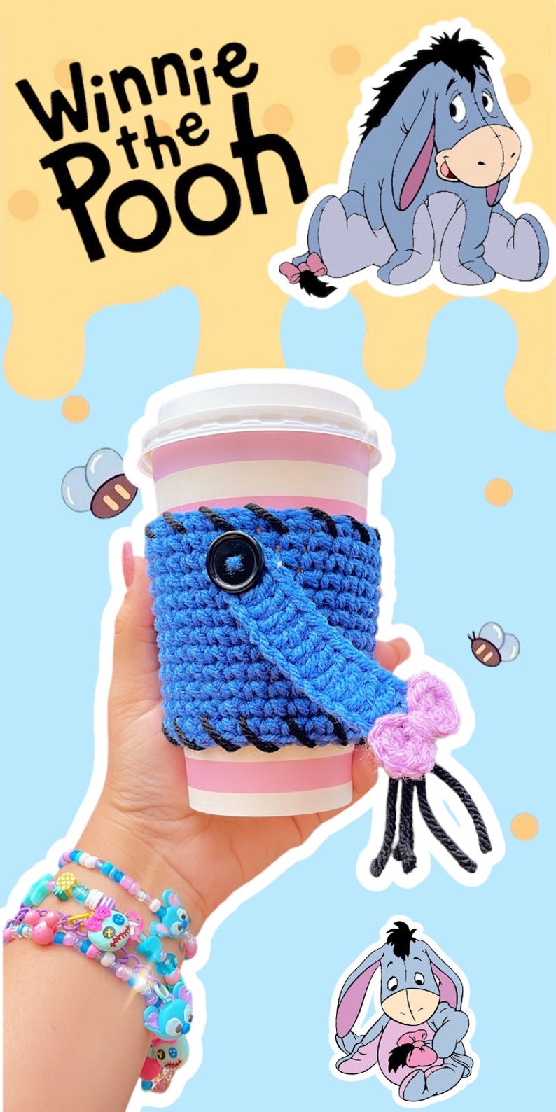 May include: A blue crocheted coffee cup sleeve with a black button and a pink tassel, held in a hand. The sleeve is wrapped around a pink and white striped cup. The image includes Winnie the Pooh characters and text.