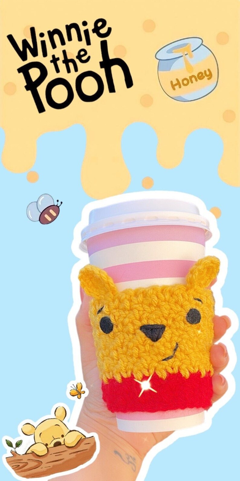 May include: A yellow and red crocheted Winnie the Pooh coffee cup cozy. The cup has a pink and white striped design. The image includes the text "Winnie the Pooh" and a honey pot illustration.