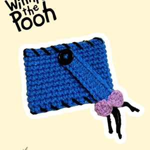 May include: A blue crocheted case with black stitching and a black button closure. A purple bow is attached to the case. The words "Winnie the Pooh" are at the top. The image also features illustrations of Eeyore, Winnie the Pooh, Piglet, and Tigger.