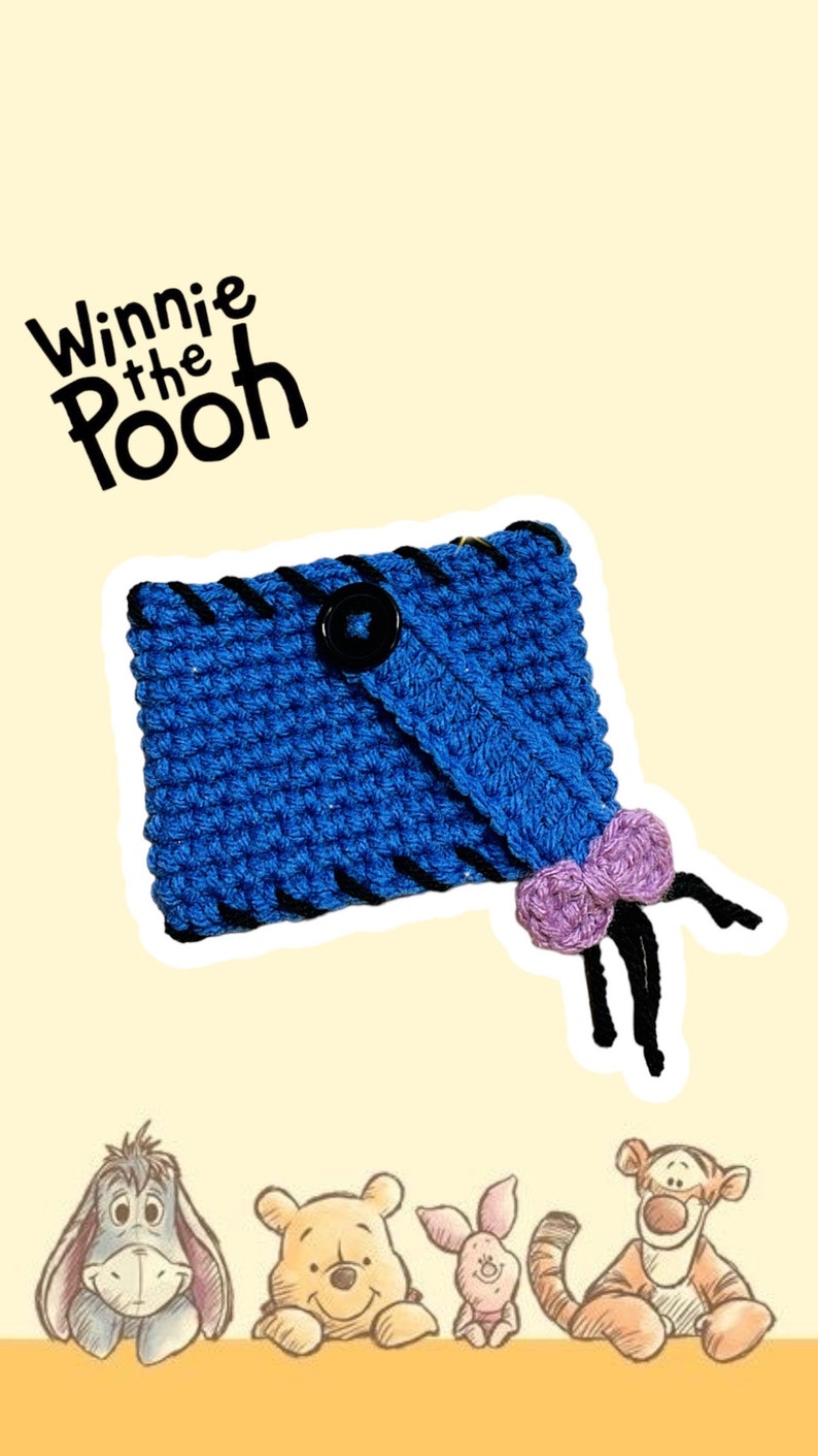 May include: A blue crocheted case with black stitching and a black button closure. A purple bow is attached to the case. The words "Winnie the Pooh" are at the top. The image also features illustrations of Eeyore, Winnie the Pooh, Piglet, and Tigger.