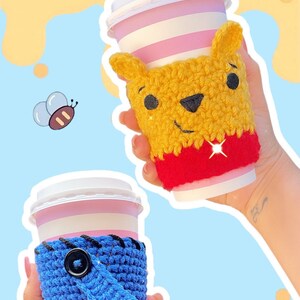May include: Two paper coffee cups with crocheted cozies. One cozy is yellow and red, resembling Winnie the Pooh. The other is blue with a black button and a pink bow. The background has Winnie the Pooh themed graphics.