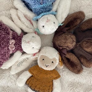 May include: A collection of four plush stuffed animals arranged in a circle. The group includes two white rabbits with embroidered names on their ears, one brown rabbit with a dark brown bow, and one white lamb wearing a mustard yellow knit sweater.