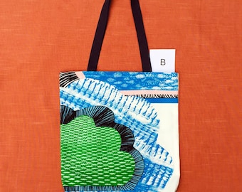 Puede incluir: Una bolsa de tela blanca con asas negras. La bolsa tiene un diseño abstracto colorido en azul, verde, negro y blanco. El diseño se asemeja a una nube o flor estilizada.