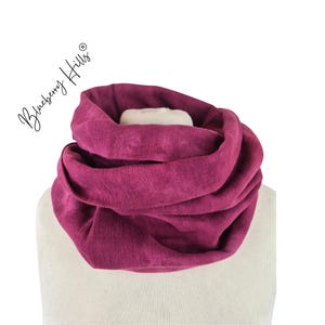 Może przedstawiać: Głęboko magenta szalik typu infinity, udrapowany na białym manekinie. Szalik wykonany jest z miękkiej, teksturowanej tkaniny i jest stylizowany w sposób okrągły. Słowa "Blueberry Hills" są napisane kursywą po lewej stronie u góry.