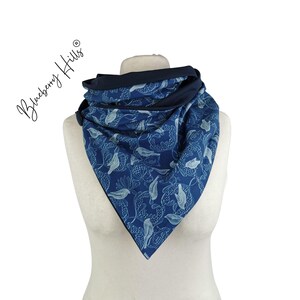 Può includere: Sciarpa blu con motivo floreale bianco e rifiniture blu scuro, esposta su un manichino. La sciarpa è triangolare e sembra essere realizzata in tessuto morbido. Il testo "Blueberry Hills" è scritto sul lato sinistro dell'immagine.