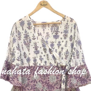 Puede incluir: Vestido cruzado con estampado floral blanco y morado, con falda escalonada y mangas acampanadas. El vestido tiene un cinturón a juego y el texto "nahata fashion shop". El vestido está colgado de una percha de madera.