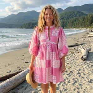 May include: A pink and white patterned dress with puffy sleeves. The dress is knee-length and has a keyhole neckline. A straw hat is held in the hand. The background features a beach, ocean, and mountains.