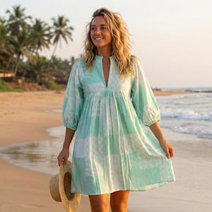 May include: A light green and white patterned dress with a v-neck and three-quarter sleeves. The dress is knee-length and is being held by a person walking on a sandy beach. A straw hat is held in the person's hand.