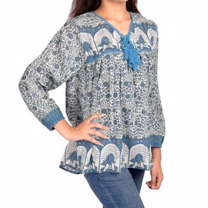 Pode incluir: Uma túnica de manga comprida com estampa floral azul e branca, com decote em V e borla azul. A túnica tem um corte folgado e é combinada com jeans azuis. A parte superior apresenta um padrão floral detalhado e acabamentos em azul.