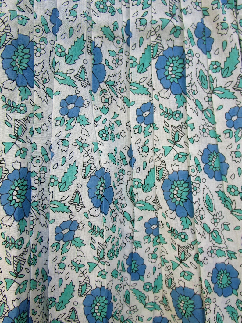 K&ouml;nnte beinhalten: Wei&szlig;er Stoff mit einem blauen und gr&uuml;nen Blumenmuster. Der Stoff ist gefaltet und hat ein zartes, vintage-m&auml;&szlig;iges Aussehen.