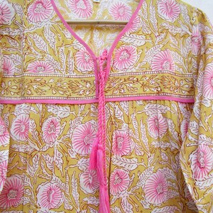 May include: A yellow and pink floral print top with a tie at the neckline and a pink tassel. The top has long sleeves and a relaxed fit.