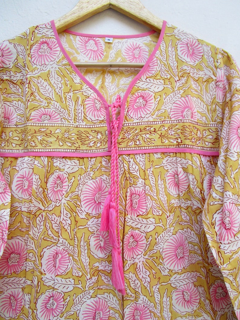 May include: A yellow and pink floral print top with a tie at the neckline and a pink tassel. The top has long sleeves and a relaxed fit.