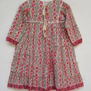 Puede incluir: Un vestido de algod&oacute;n blanco con estampado floral rosa y verde. El vestido tiene un escote redondo, mangas largas y una falda fruncida. El vestido se ata en la espalda con una borla blanca.